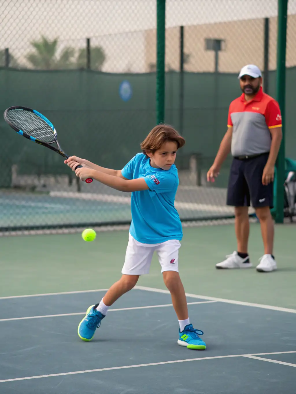 A group of young children participating in a beginner tennis lesson on a sunny outdoor court at Tennis Club de Houilles, with a coach demonstrating proper form.